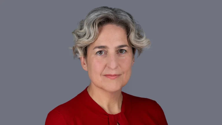 A woman with short wavy grey hair and a red shirt against a grey background.