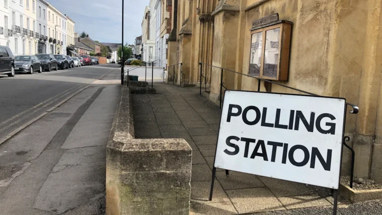 A white 'Polling Station' sign stands on a pavement outside a traditional building in a residential neighborhood.