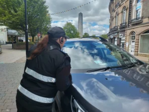 A parking enforcement officer in a black reflective vest looks into a car on a street.