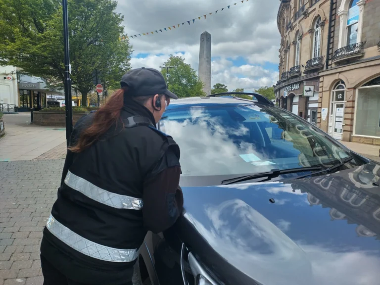 A parking enforcement officer in a black reflective vest looks into a car on a street.