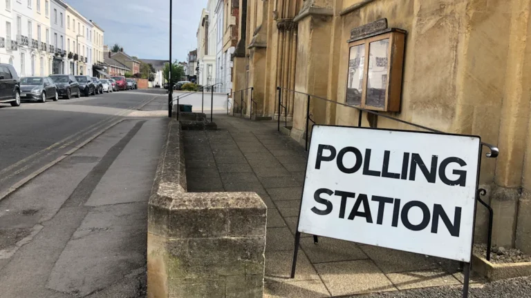 A white sign with black text reading 'POLLING STATION' stands on a sidewalk outside a stone building in a residential area.