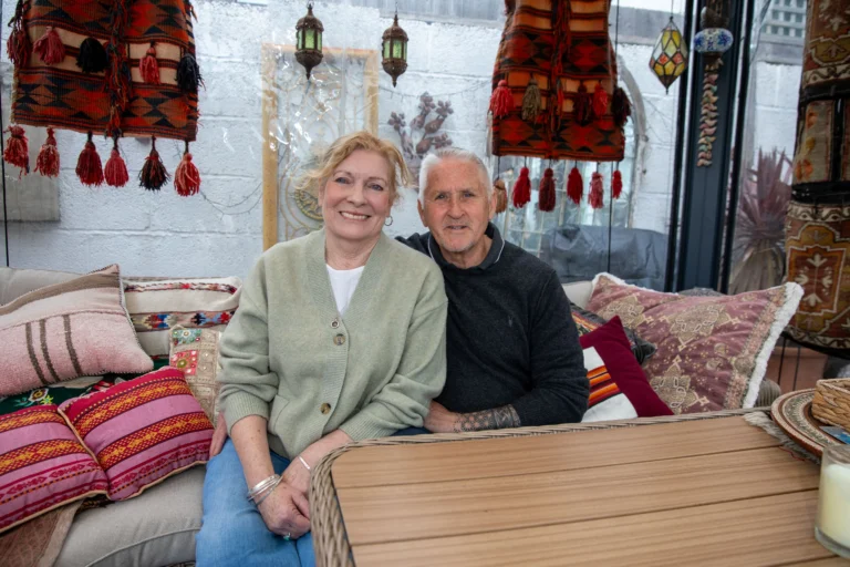 Senior man and woman sitting together on a patio decorated with colorful rugs and pillows.