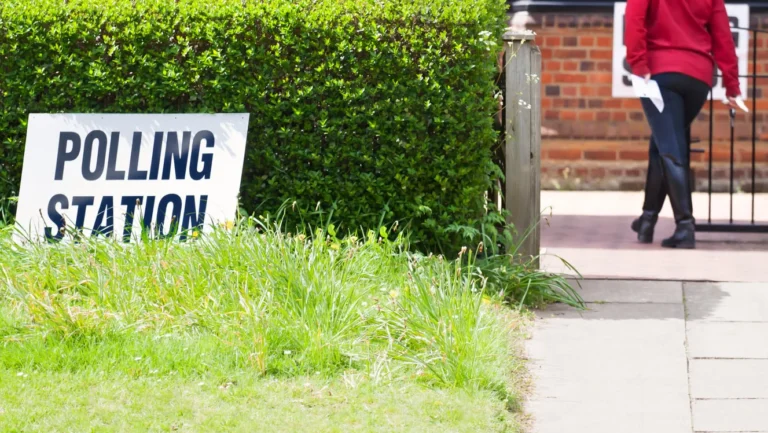 A white sign reading "POLLING STATION" positioned in front of a lush green hedge.