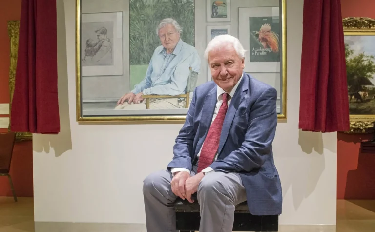 David Attenborough sits in front of a large painted portrait of himself in an art gallery.