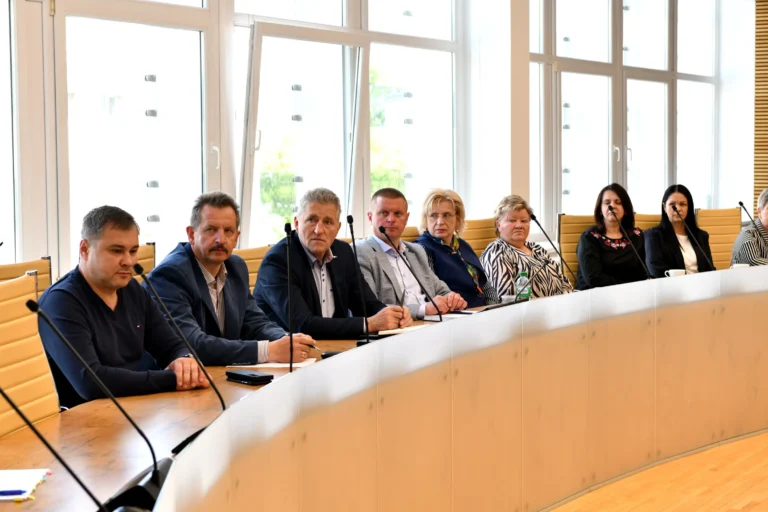 A group of people sitting at a long curved table with microphones in front of windows.