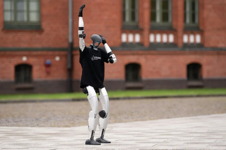 A humanoid robot in a black t-shirt posing with one arm raised outdoors.