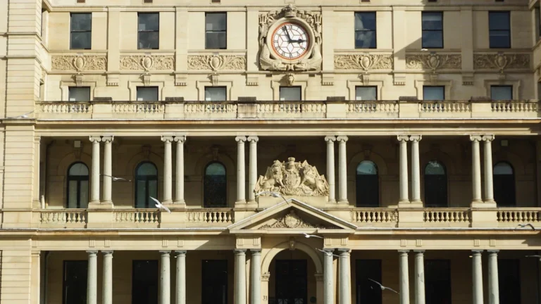 Ornate stone building facade with Neoclassical columns and a large central clock under bright sunlight.