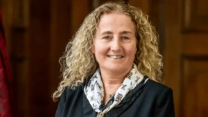 A smiling woman with long blonde curly hair wearing a black blazer and patterned scarf.