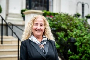 Smiling woman with long curly blonde hair wearing a black blazer and patterned scarf outdoors.