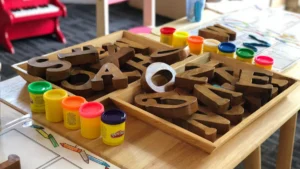 Wooden alphabet letters and colorful play dough containers on a nursery school table with a toy piano.