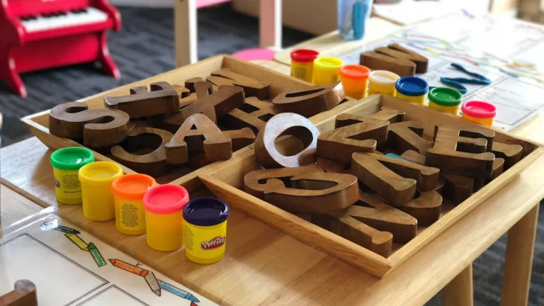 Wooden alphabet letters and colorful play dough containers on a nursery school table with a toy piano.