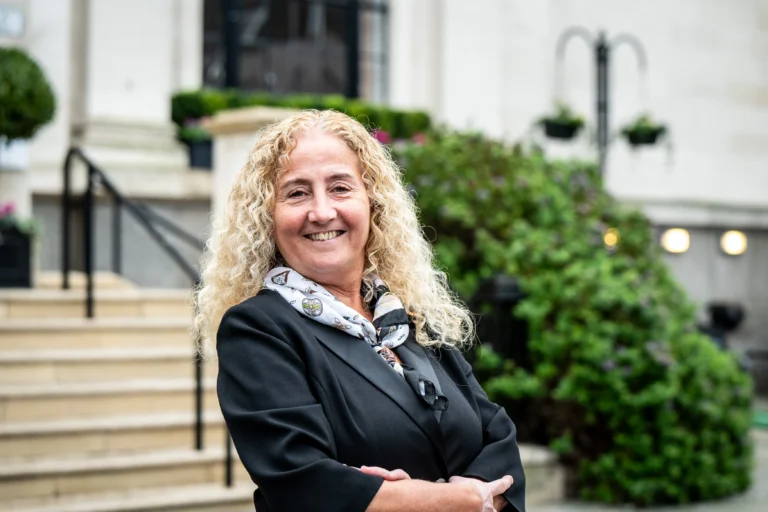 A smiling woman with long curly blonde hair wearing a black blazer and patterned scarf.