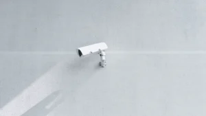 A white modern box security camera mounted on a plain, light-colored exterior wall.