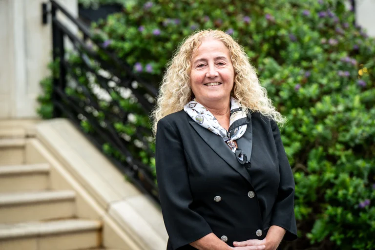 Smiling woman with curly blonde hair wearing a black blazer and patterned scarf standing outdoors.