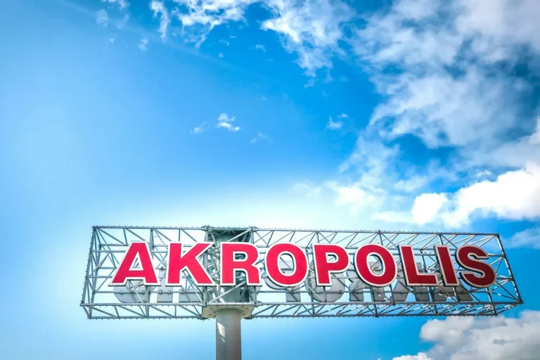 Large red AKROPOLIS letters on a metal frame against a bright blue sky with clouds.