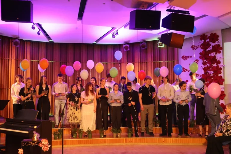 Group of students holding colorful balloons on a stage with purple lighting and wooden backdrops.