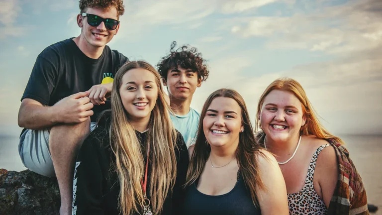 A diverse group of five smiling teenagers and young adults posing together outdoors during summer.