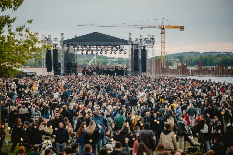 Large crowd gathered before an outdoor music stage on a riverbank with a construction crane.