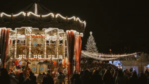 A glowing vintage carousel and tall Christmas tree at an outdoor community festival at night.