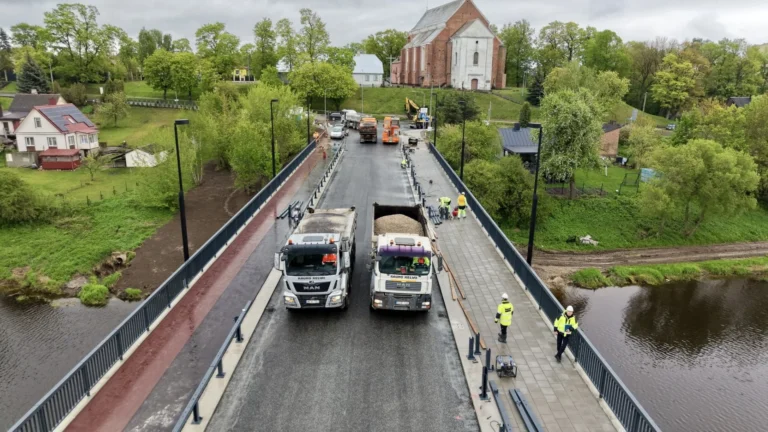Two white dump trucks parked on a newly paved bridge with workers in high-visibility vests.