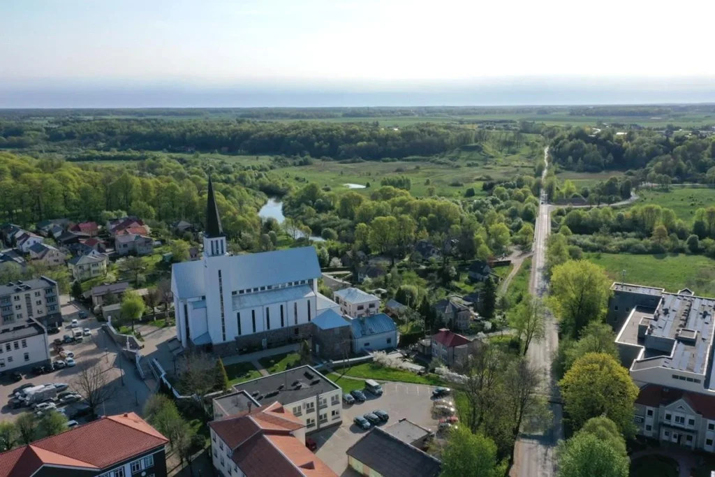 Aerial view of a white church with a black spire in a green landscape with a river.
