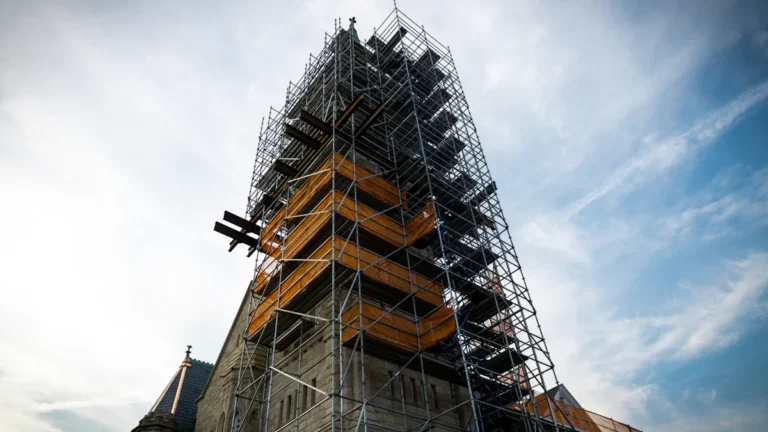 A tall church tower completely encased in metal scaffolding against a cloudy sky during a major restoration project.