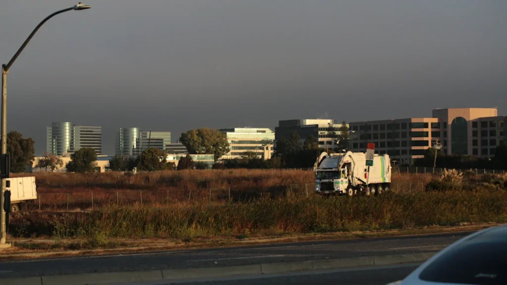 A white waste collection truck drives along a road past a field and commercial buildings.