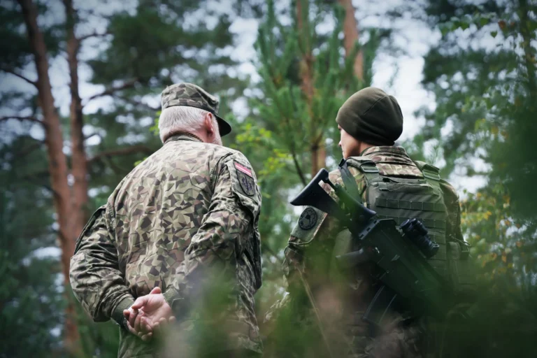 Two soldiers in camouflage uniforms stand in a forest, one carrying a rifle on their back.