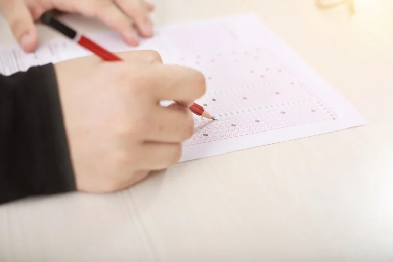 Hands using a red pencil to mark answers on a multiple-choice optical mark recognition sheet.