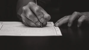 A close-up black and white photograph of a hand signing a formal document, representing the signature collection process for Latvia's pension reform.