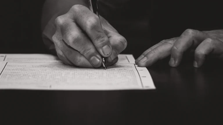 A close-up black and white photograph of a hand signing a formal document, representing the signature collection process for Latvia's pension reform.