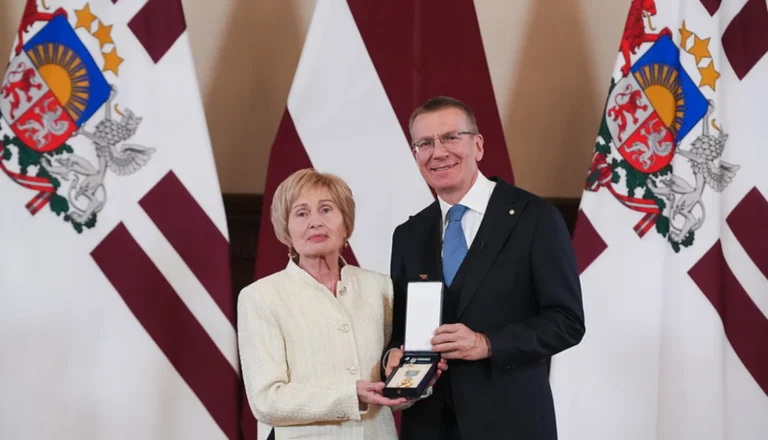 A man and woman holding an open medal box in front of three Latvian flags.