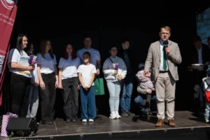 A man in a checkered jacket speaks into a microphone while people stand on a stage.