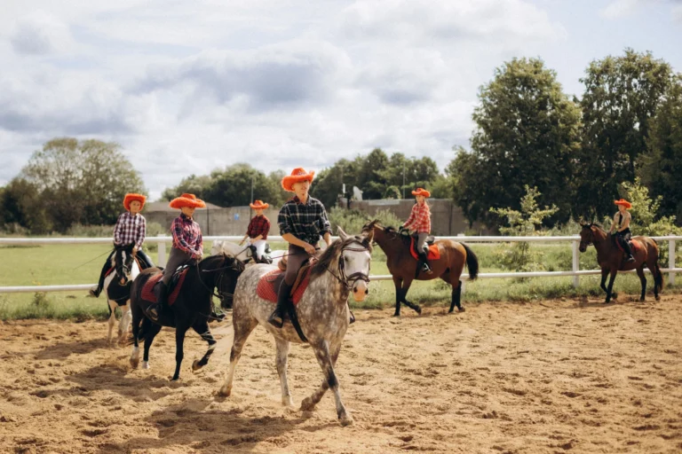 Six young riders in orange hats and plaid shirts ride horses through a sandy arena.