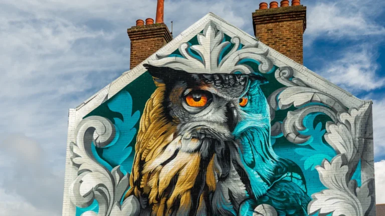 A vibrant mural of a large owl with intense orange eyes on a UK brick building.