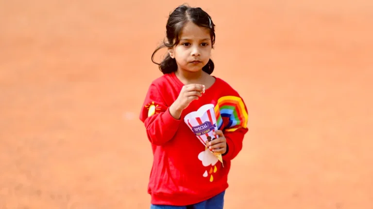 A young girl in a red sweatshirt holding a paper cone of snacks outdoors.