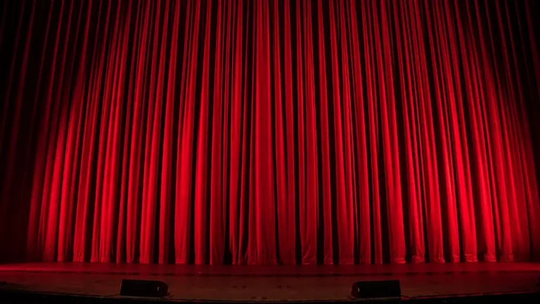 A large, pleated red velvet stage curtain illuminated by spotlights in a theater.