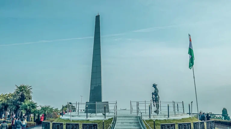 A bronze soldier statue stands before a tall dark obelisk at a war memorial under a clear blue sky.