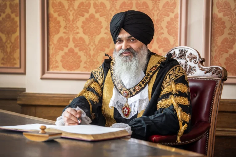 A smiling man in ceremonial robes and a turban signing an official document at a desk.