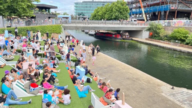 A vibrant crowd of people enjoying a sunny day at a riverside festival with seating areas and a bridge.