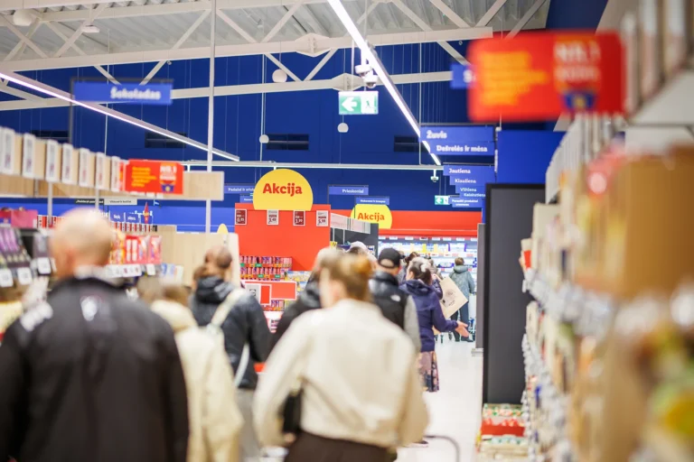 Shoppers walk down a supermarket aisle past yellow sale signs under bright blue ceilings and lights.