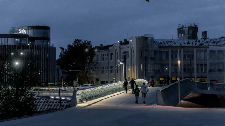 People walking across a modern illuminated bridge at night in Kaunas, Lithuania, with city buildings behind.