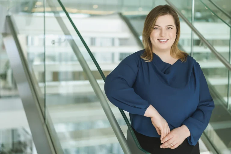 A smiling woman with light brown hair wearing a blue blouse leans against a glass staircase railing.