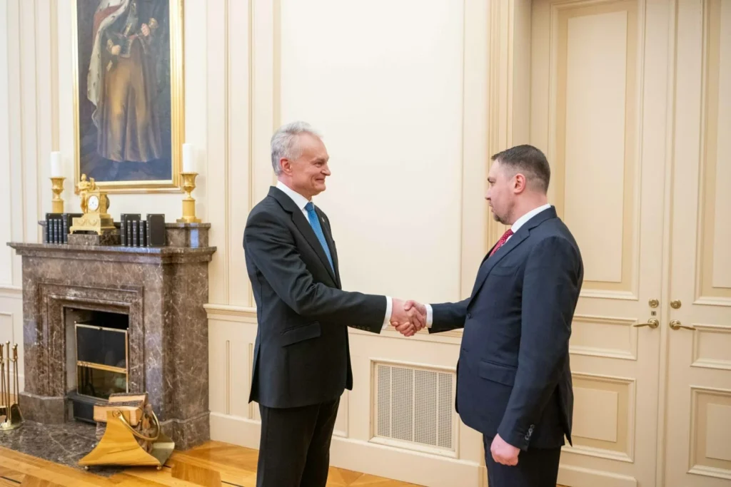 Two men in formal suits shake hands in a room featuring a marble fireplace and painting.