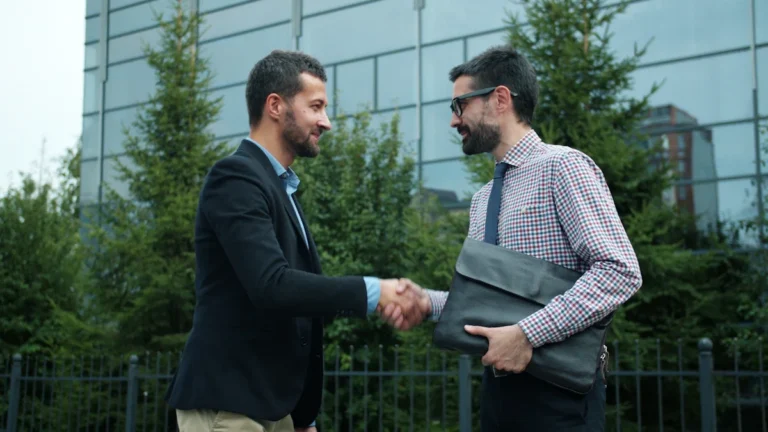 Two men in business suits shaking hands outside a modern glass office building.