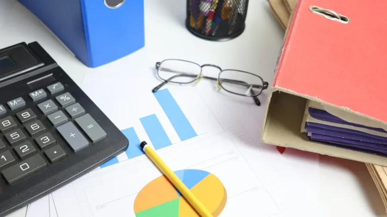 A desk viewed from above with a calculator, pie charts, bar graphs, binders, and reading glasses.