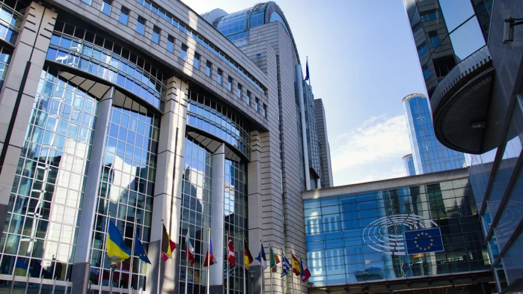 The modern glass architecture of the European Parliament building with member state flags flying in front.