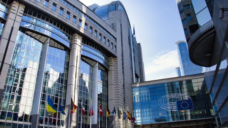 The modern glass architecture of the European Parliament building with member state flags flying in front.