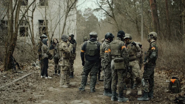 A group of individuals in tactical gear and camouflage participating in a civil defense training exercise in a wooded area near a ruined building.