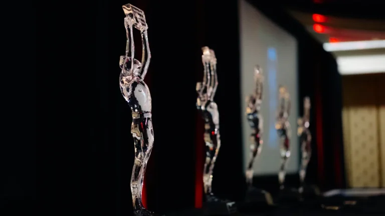 Clear crystal trophies shaped like human figures lined up on a dark stage under spotlighting.